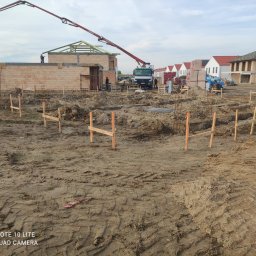Usługi Ogólnobudowlane Maciej Bąk - Przygotowanie terenu pod budowę domu z widocznymi wykopami, drewnianymi szalunkami, zbrojeniami, częściowo wzniesionymi ścianami z cegły oraz pompą do betonu w tle.
