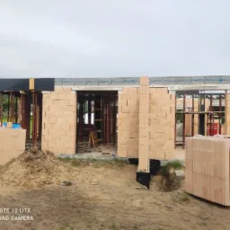 Wznoszenie ścian domu z bloczków ceramicznych Porotherm, widoczne szalunki, podpory i wiązania stropowe, paleta bloczków na pierwszym planie, etap budowy w stanie surowym otwartym.