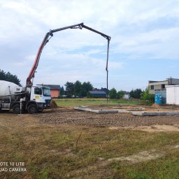 Usługi Ogólnobudowlane Maciej Bąk - Betonomieszarka z wysięgnikiem pompuje beton do przygotowanych szalunków fundamentowych na placu budowy. Widoczne zbrojenia i ubity grunt.