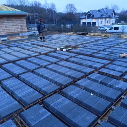 Usługi Ogólnobudowlane Maciej Bąk - Budowa stropu Teriva: pracownik stoi na ułożonych bloczkach stropowych, widoczne zbrojenie, w tle budynek w stanie surowym otwartym i dom jednorodzinny.