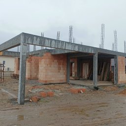 Usługi Ogólnobudowlane Maciej Bąk - Wznoszona konstrukcja domu z czerwonej cegły, betonowe słupy i nadproża, wystające pręty zbrojeniowe, widoczne ślady błota na placu budowy, pochmurne niebo w tle.