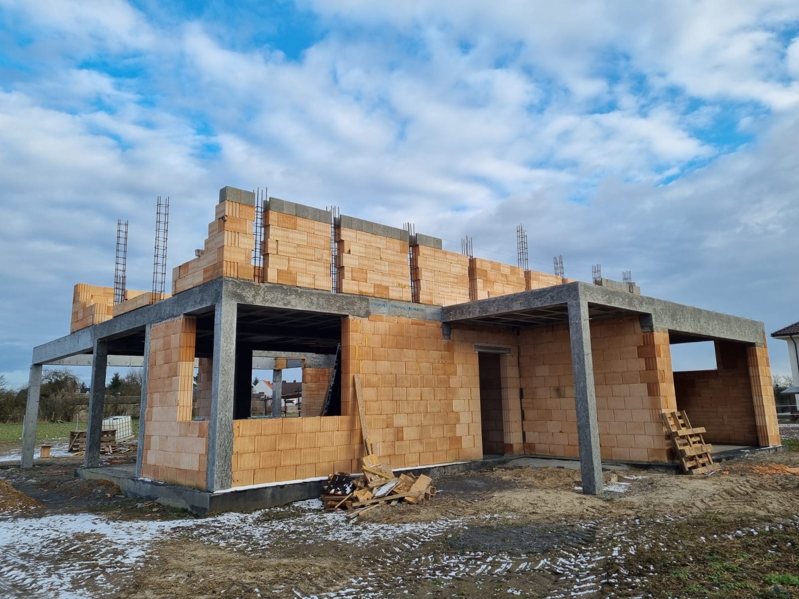 Surowy stan domu jednorodzinnego w trakcie budowy, widoczne ściany z cegły ceramicznej Porotherm, betonowe nadproża i słupy, zbrojenie pionowe, częściowo ośnieżony teren.