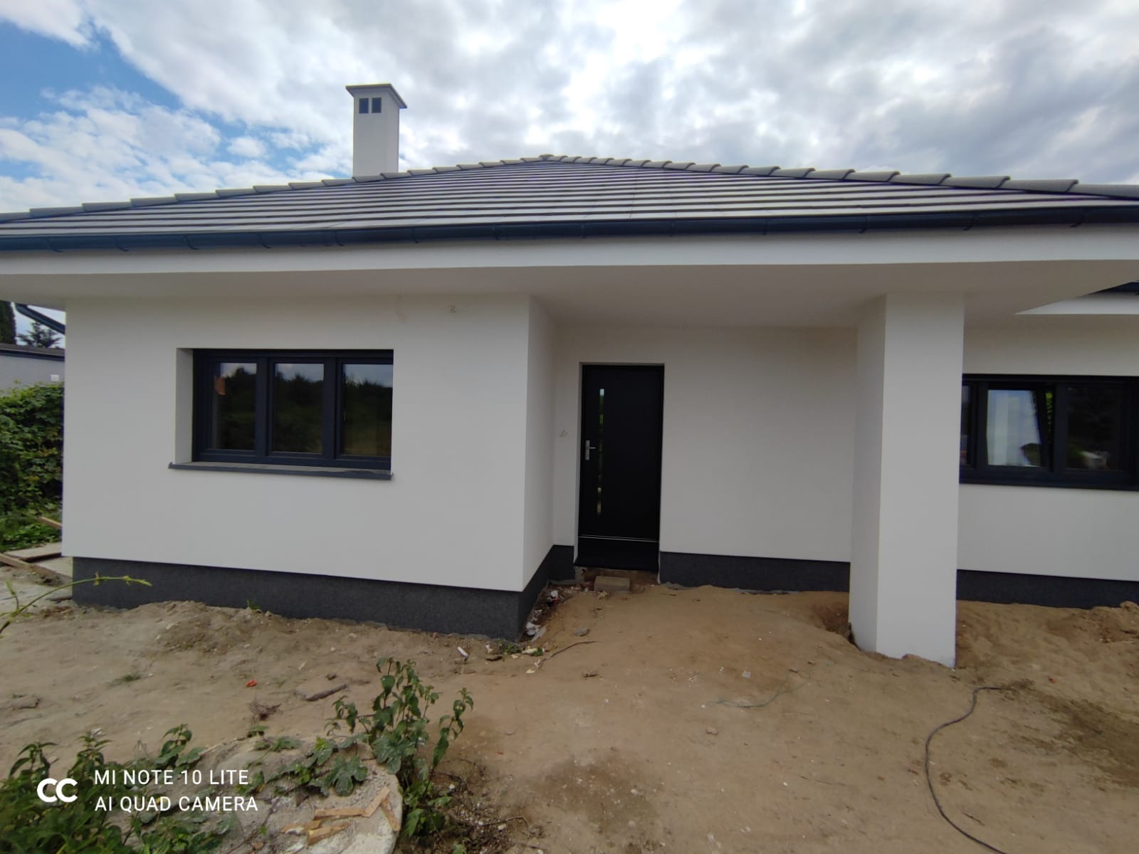 Nowoczesny dom jednorodzinny z białymi ścianami i grafitową stolarką okienną oraz dachem z szarej dachówki, widoczny fragment elewacji i teren wokół domu w trakcie prac wykończeniowych.