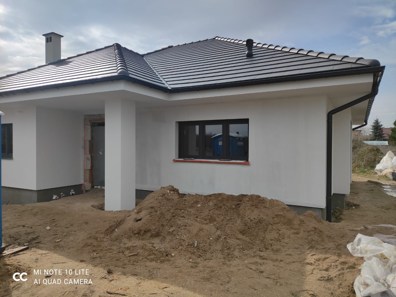 Nowoczesny dom jednorodzinny z ciemną dachówką i białymi ścianami, w trakcie wykończenia terenu wokół budynku, widoczna sterta ziemi i niezabudowany otwór drzwiowy.
