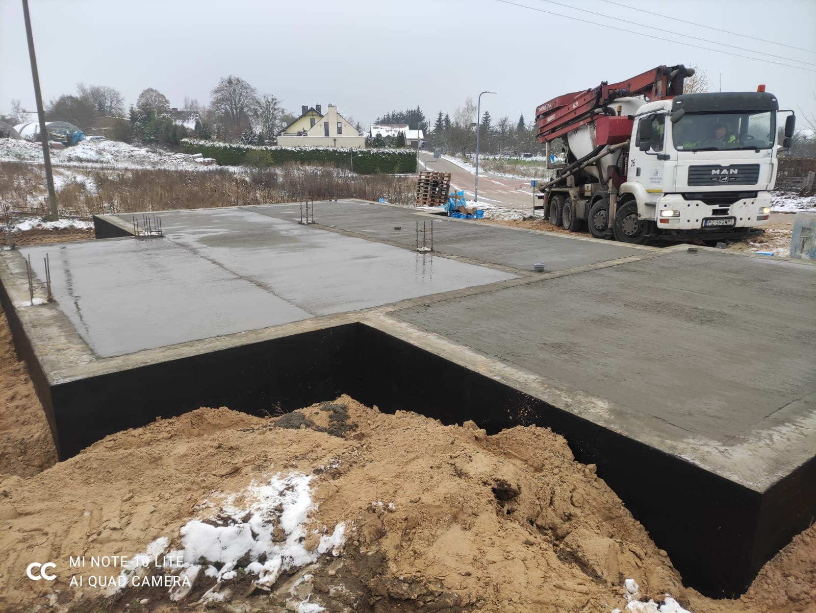 Wylany beton na płycie fundamentowej z wystającymi prętami zbrojeniowymi, obok stoi betonomieszarka MAN na tle zimowego krajobrazu.