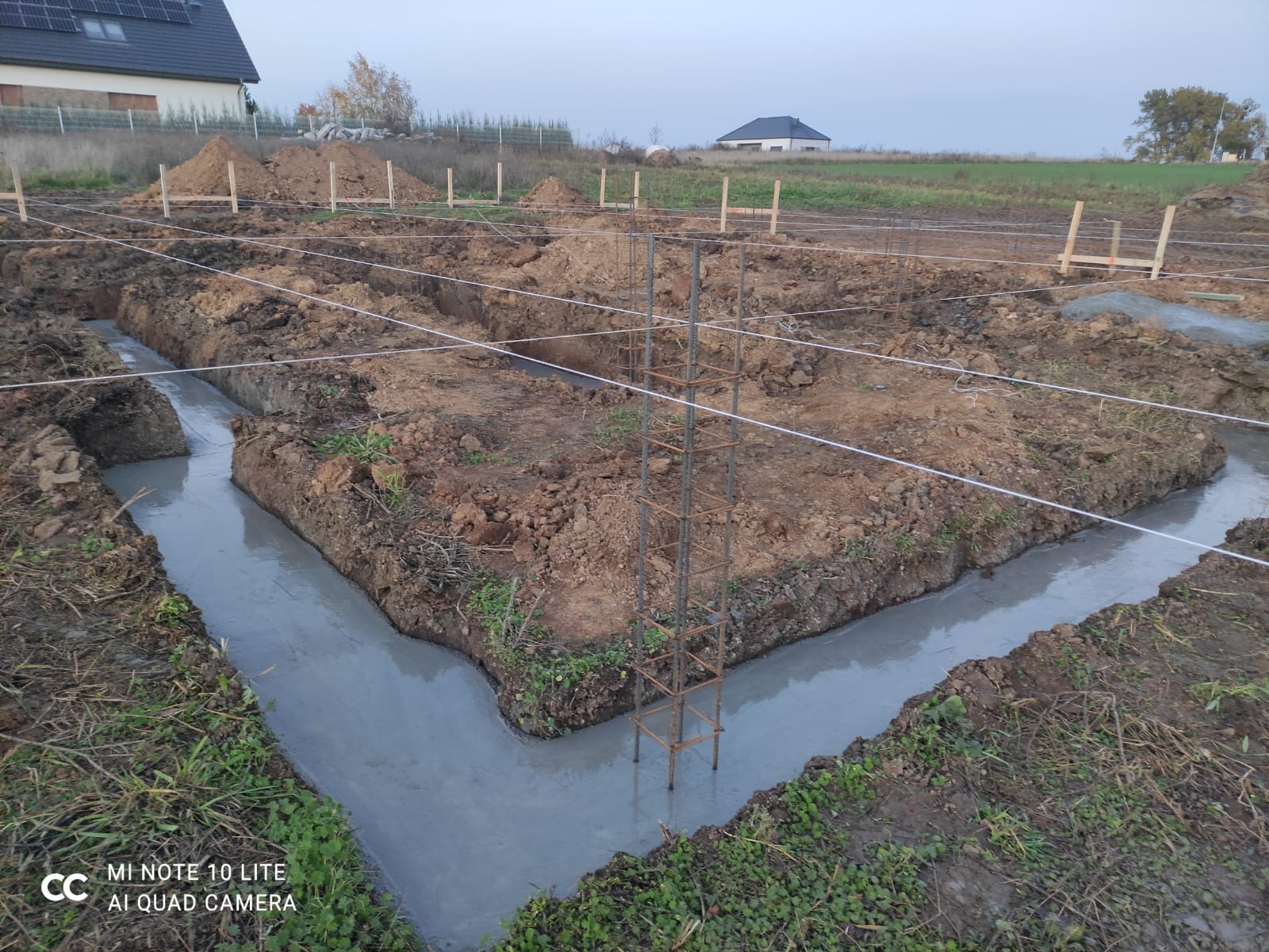 Wylane fundamenty w wykopie, zbrojenie stalowe w narożniku, widoczne sznurki wyznaczające granice, w tle ziemia i domy.