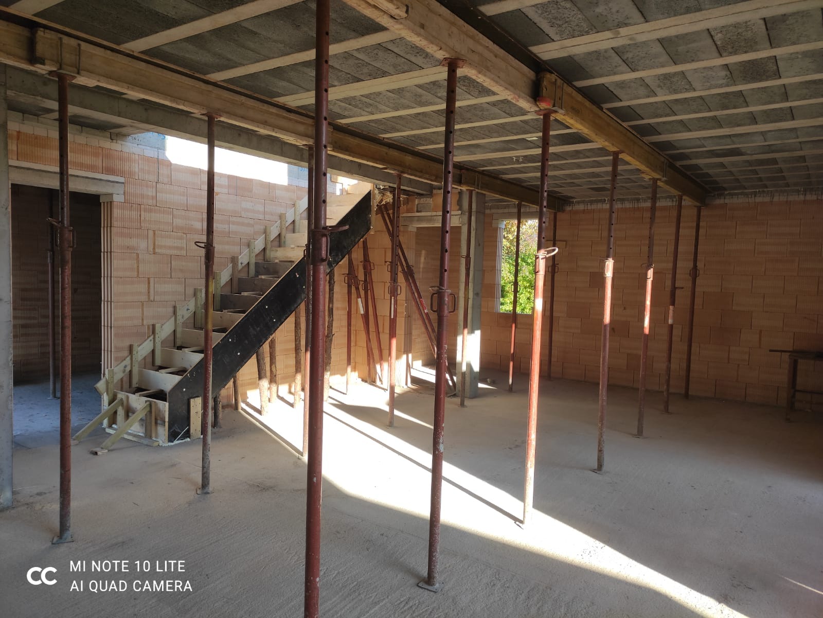 Wnętrze w trakcie budowy domu z cegły, widoczne ściany, drewniane szalunki stropu podparte metalowymi stemplami, tymczasowe drewniane schody.