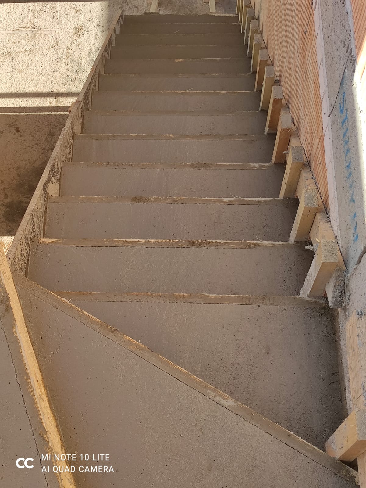 Surowe betonowe schody w trakcie budowy, z widocznym drewnianym szalunkiem i fragmentem murowanej ściany w tle.