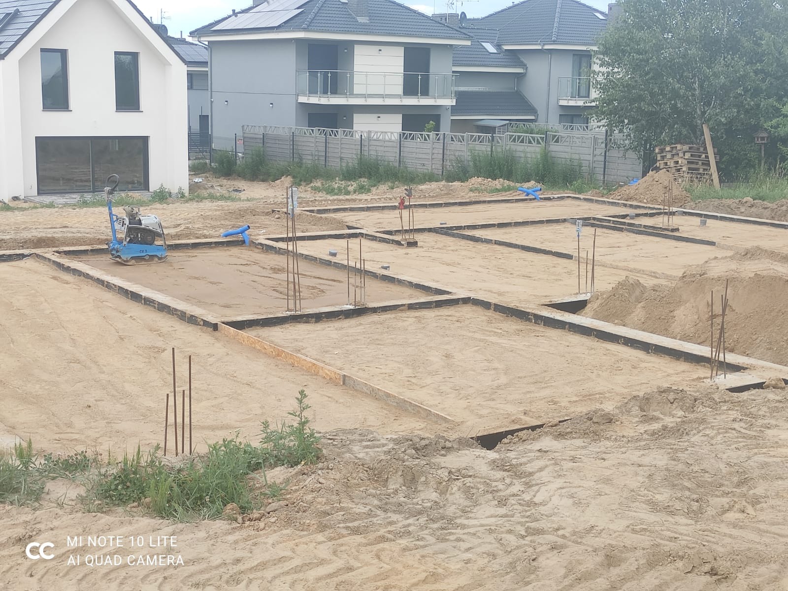 Przygotowanie terenu pod budowę: wykopane fundamenty z szalunkiem, wystające pręty zbrojeniowe, ubijarka wibracyjna w tle, nowe domy jednorodzinne w tle.