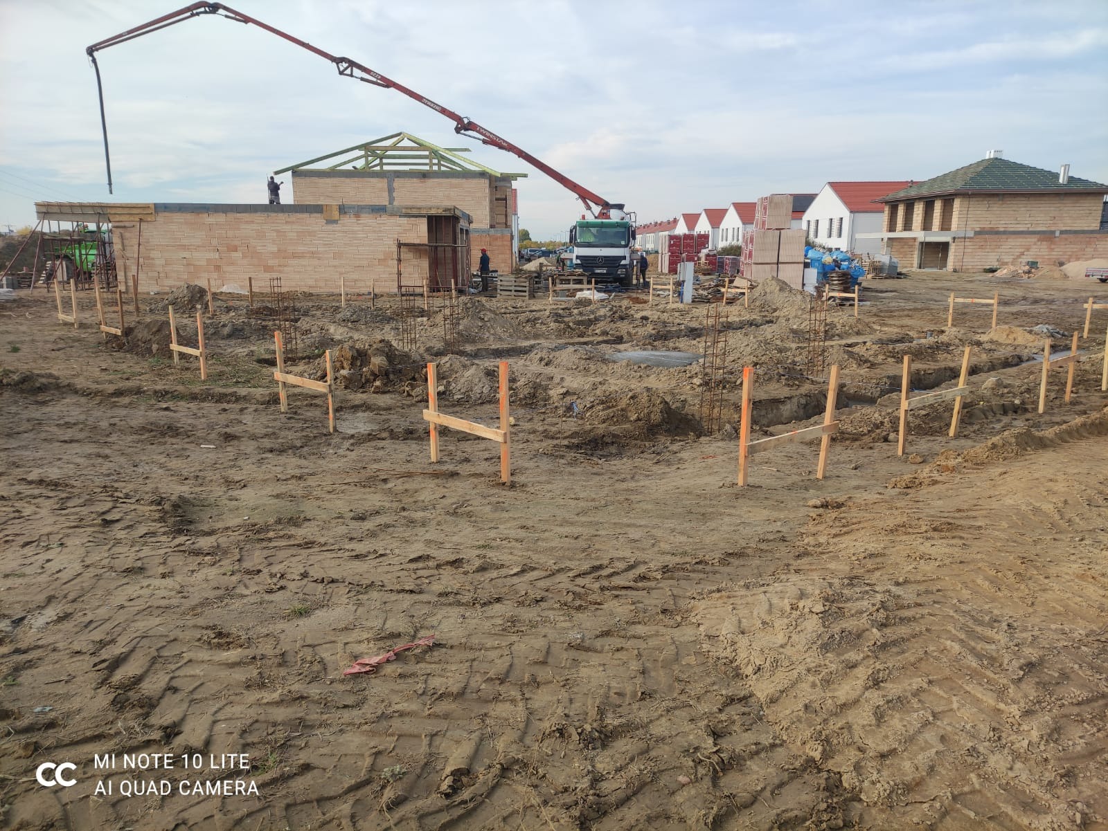Przygotowanie terenu pod budowę domu z widocznymi wykopami, drewnianymi szalunkami, zbrojeniami, częściowo wzniesionymi ścianami z cegły oraz pompą do betonu w tle.