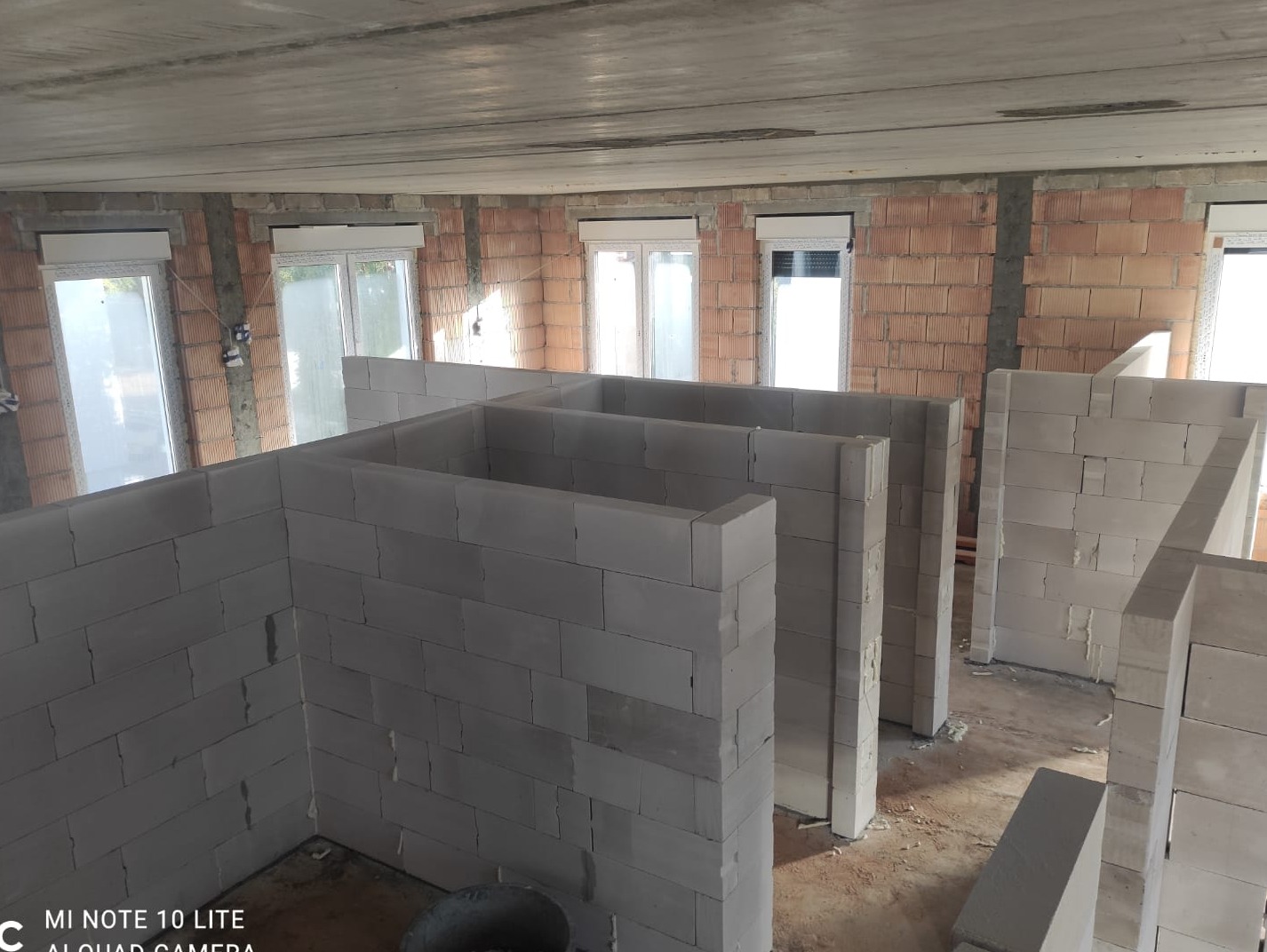 Wznoszenie ścian działowych z bloczków betonu komórkowego w surowym stanie, widoczne okna i ceglany mur w tle.