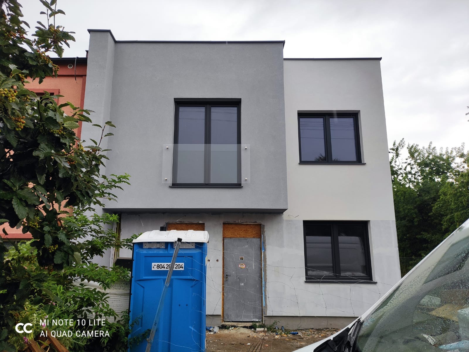 Dwukondygnacyjny dom murowany w trakcie budowy, z widocznymi oknami w czarnych ramach, szarą elewacją, tymczasowymi drzwiami wejściowymi, niebieską toaletą przenośną i elementami rusztowania.