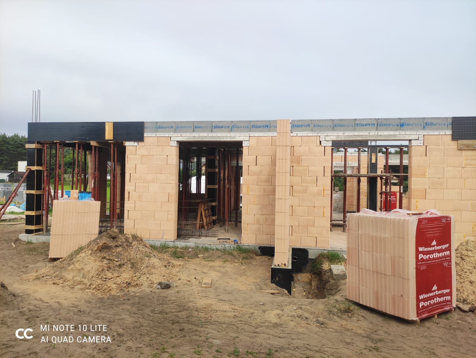 Wznoszenie ścian domu z bloczków ceramicznych Porotherm, widoczne szalunki, podpory i wiązania stropowe, paleta bloczków na pierwszym planie, etap budowy w stanie surowym otwartym.