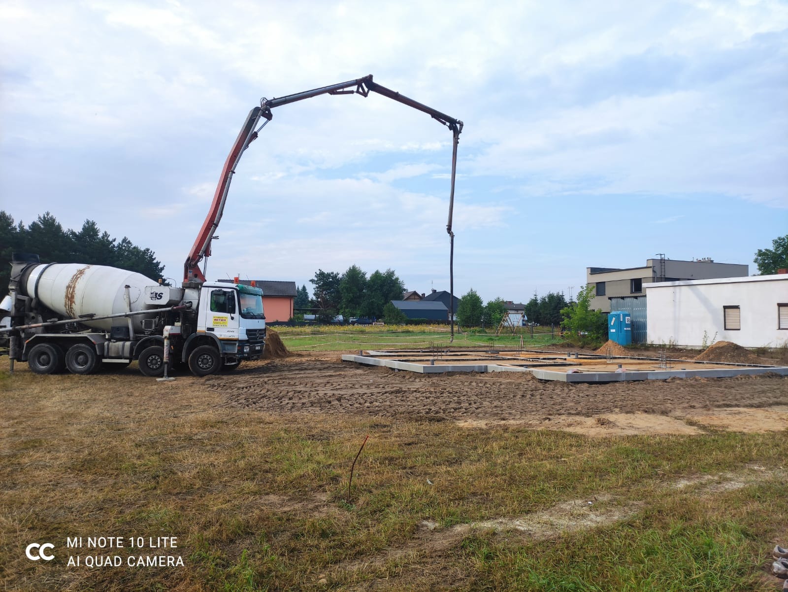 Betonomieszarka z wysięgnikiem pompuje beton do przygotowanych szalunków fundamentowych na placu budowy. Widoczne zbrojenia i ubity grunt.