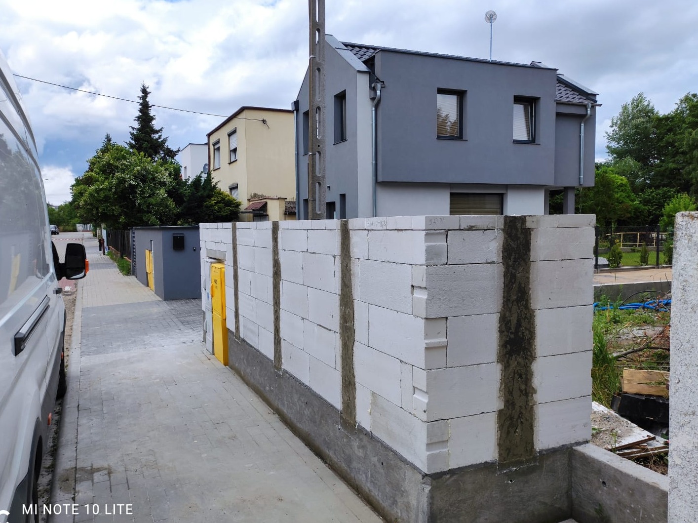 Wznoszenie muru oporowego z białych bloczków betonowych na betonowym fundamencie, widoczne spoiny pionowe wypełnione zaprawą, w tle zabudowa jednorodzinna i częściowo widoczny samochód dostawczy.
