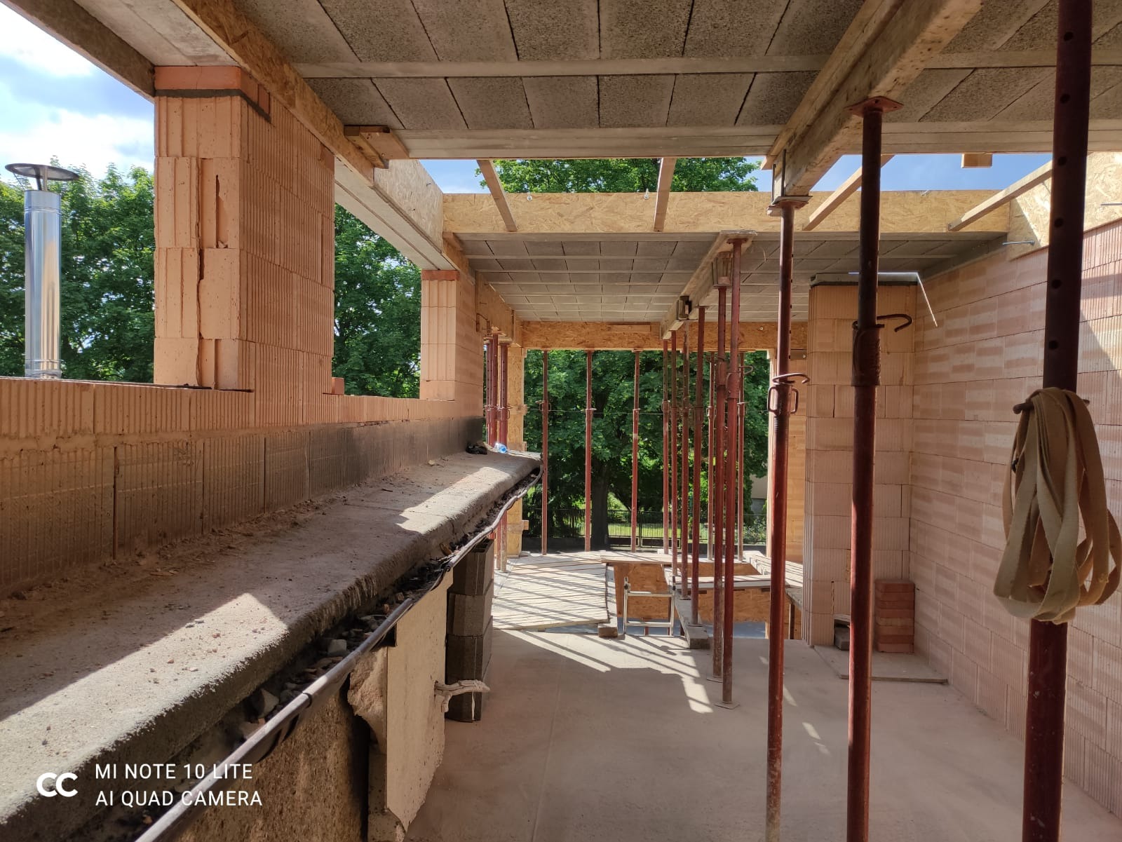 Wnętrze domu w trakcie budowy: ściany z czerwonej cegły, stalowe podpory stropu, widok na zewnątrz przez otwór okienny z kominem.