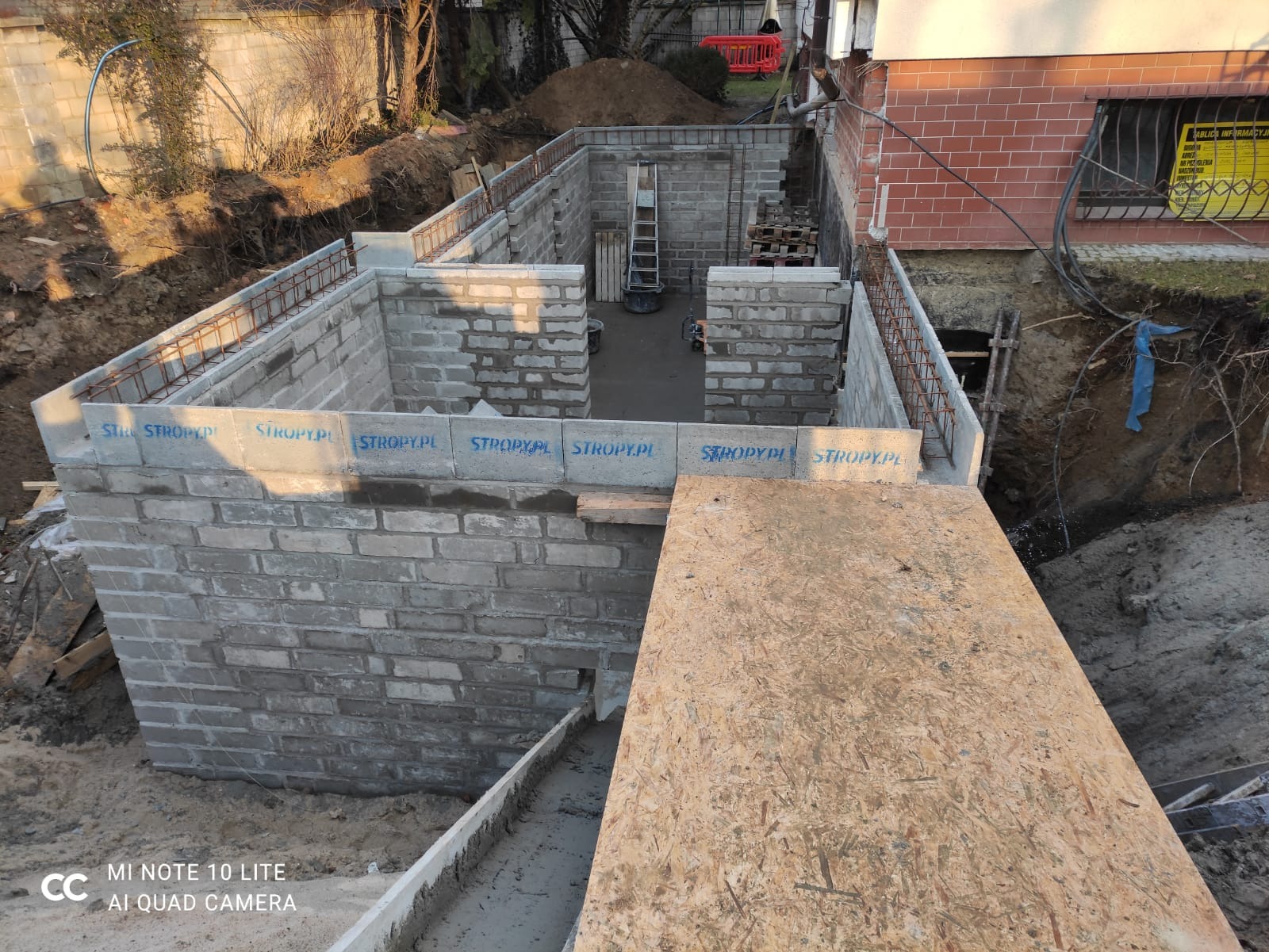 Wykop pod fundamenty domu z częściowo wymurowanymi ścianami z bloczków betonowych, widoczne zbrojenie, szalunki i tymczasowe przejście z płyty OSB.