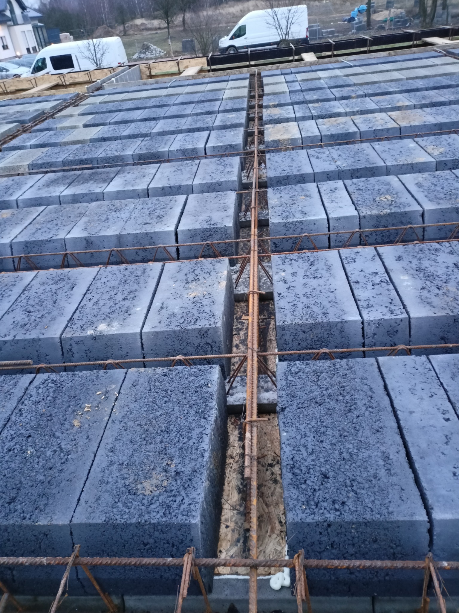 Wiązanie zbrojeniowe pomiędzy bloczkami betonowymi na fundamencie w trakcie budowy, widok z góry, w tle zabudowa jednorodzinna i biały samochód dostawczy.
