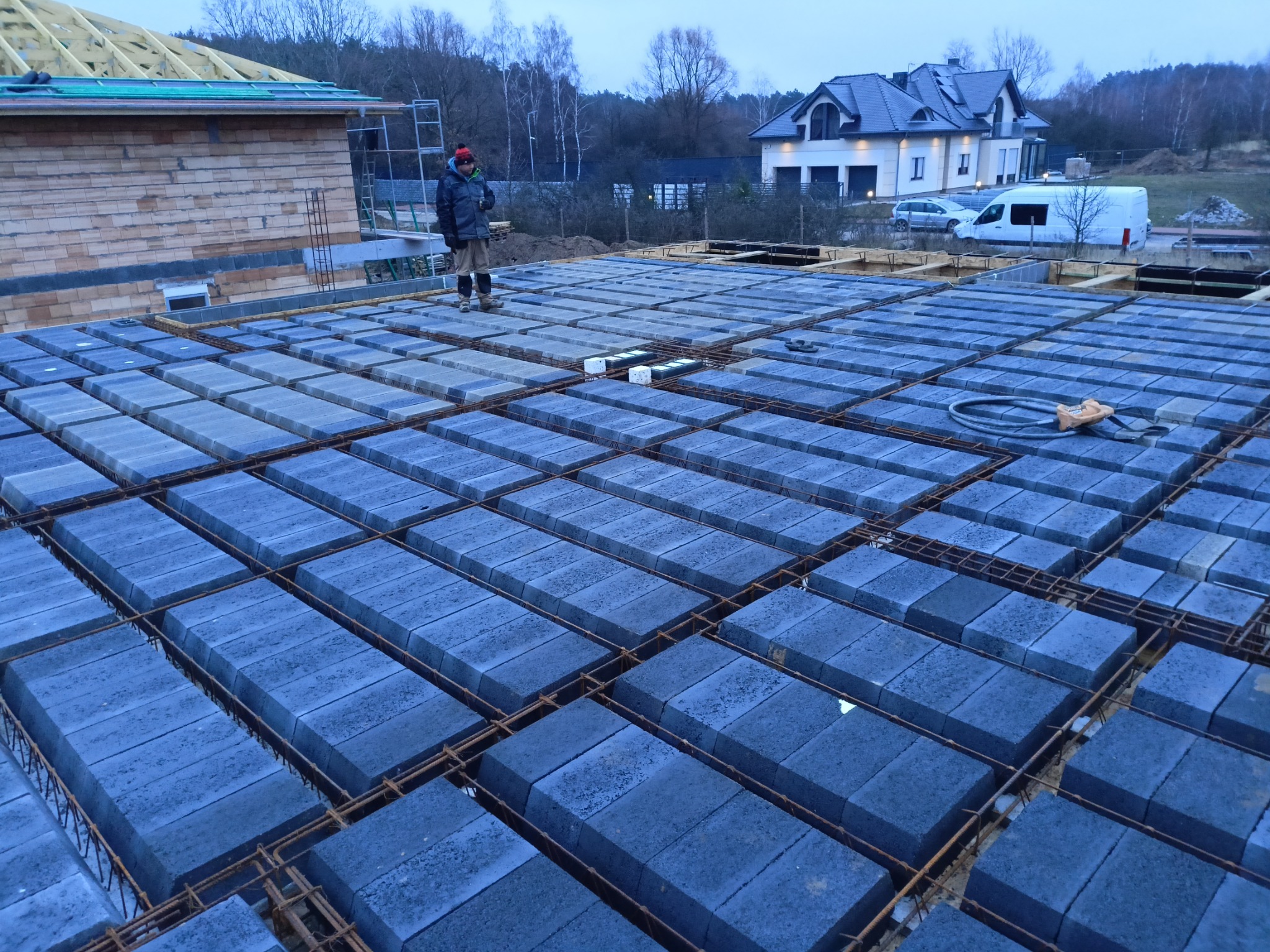 Budowa stropu Teriva: pracownik stoi na ułożonych bloczkach stropowych, widoczne zbrojenie, w tle budynek w stanie surowym otwartym i dom jednorodzinny.