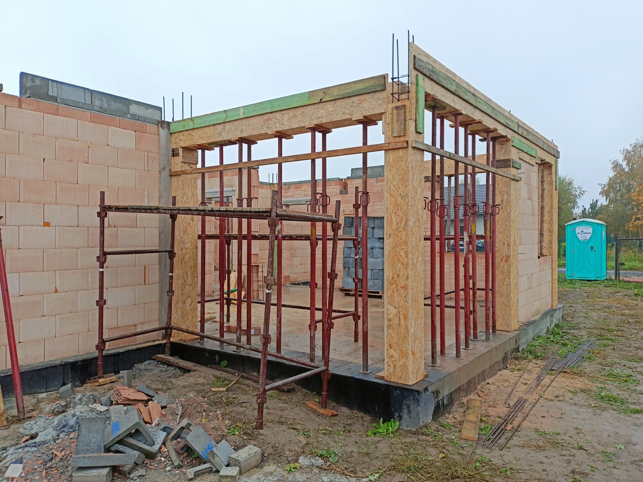 Wznoszenie ścian domu z bloczków ceramicznych, widoczne szalunki drewniane i stalowe podpory, teren budowy z toaletą przenośną w tle.