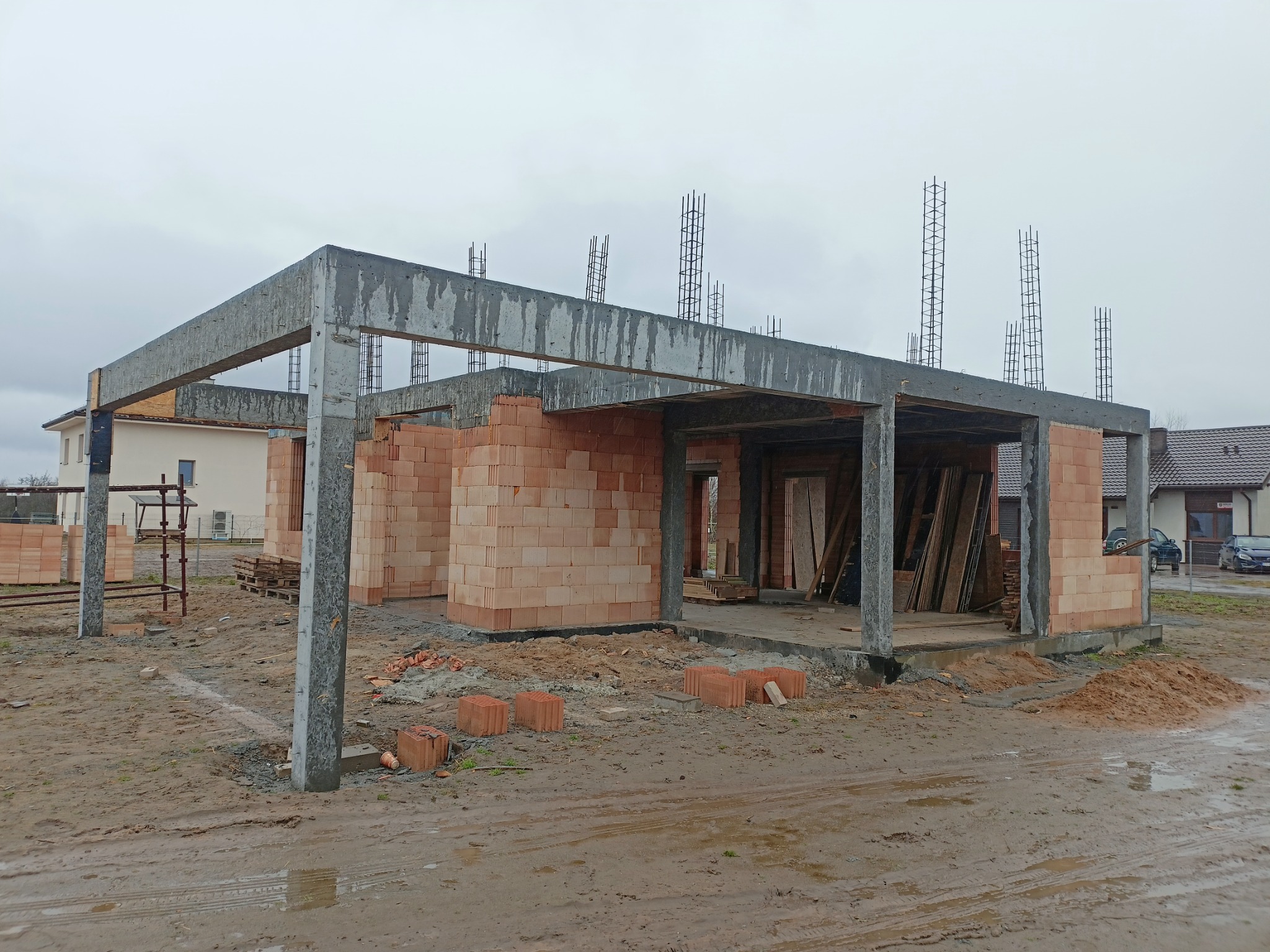 Wznoszona konstrukcja domu z czerwonej cegły, betonowe słupy i nadproża, wystające pręty zbrojeniowe, widoczne ślady błota na placu budowy, pochmurne niebo w tle.