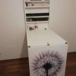 Biała składana szafka z półkami na lakiery do paznokci, ozdobiona motywem dmuchawca, zamontowana na ścianie, na podłodze panele.