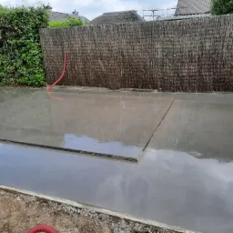 Świeżo wylana, mokra posadzka betonowa w Brzesku, z widocznymi dylatacjami i odbiciem nieba, otoczona drewnianym szalunkiem i ogrodzeniem z wikliny.