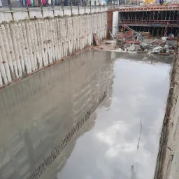 Wykop fundamentowy zabezpieczony ścianką berlińską, zalany świeżym betonem, w tle widoczne budynki mieszkalne i elementy konstrukcji szalunkowej.