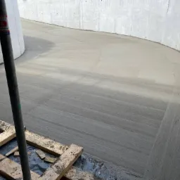 Świeżo wylana, gładka, szara betonowa posadzka w zakrzywionym pomieszczeniu przemysłowym, wsparta metalową podporą, z widocznymi deskami szalunkowymi w rogu.