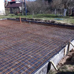 Zbrojenie płyty fundamentowej ułożone na czarnej folii, widoczne szalunki z desek i podpory, w tle trawa i drzewa