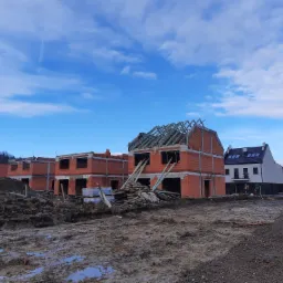 Wznoszone domy z czerwonej cegły w stanie surowym otwartym, widoczne drewniane elementy konstrukcji dachu, teren budowy z błotem i kałużami, w tle gotowy dom i pracownicy w odblaskowych kamizelkach.