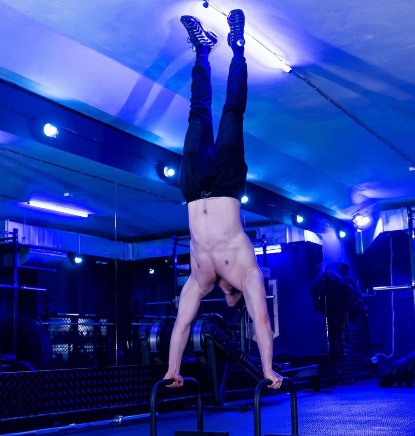 Mężczyzna bez koszulki wykonuje stójkę na poręczach w siłowni oświetlonej niebieskim światłem.