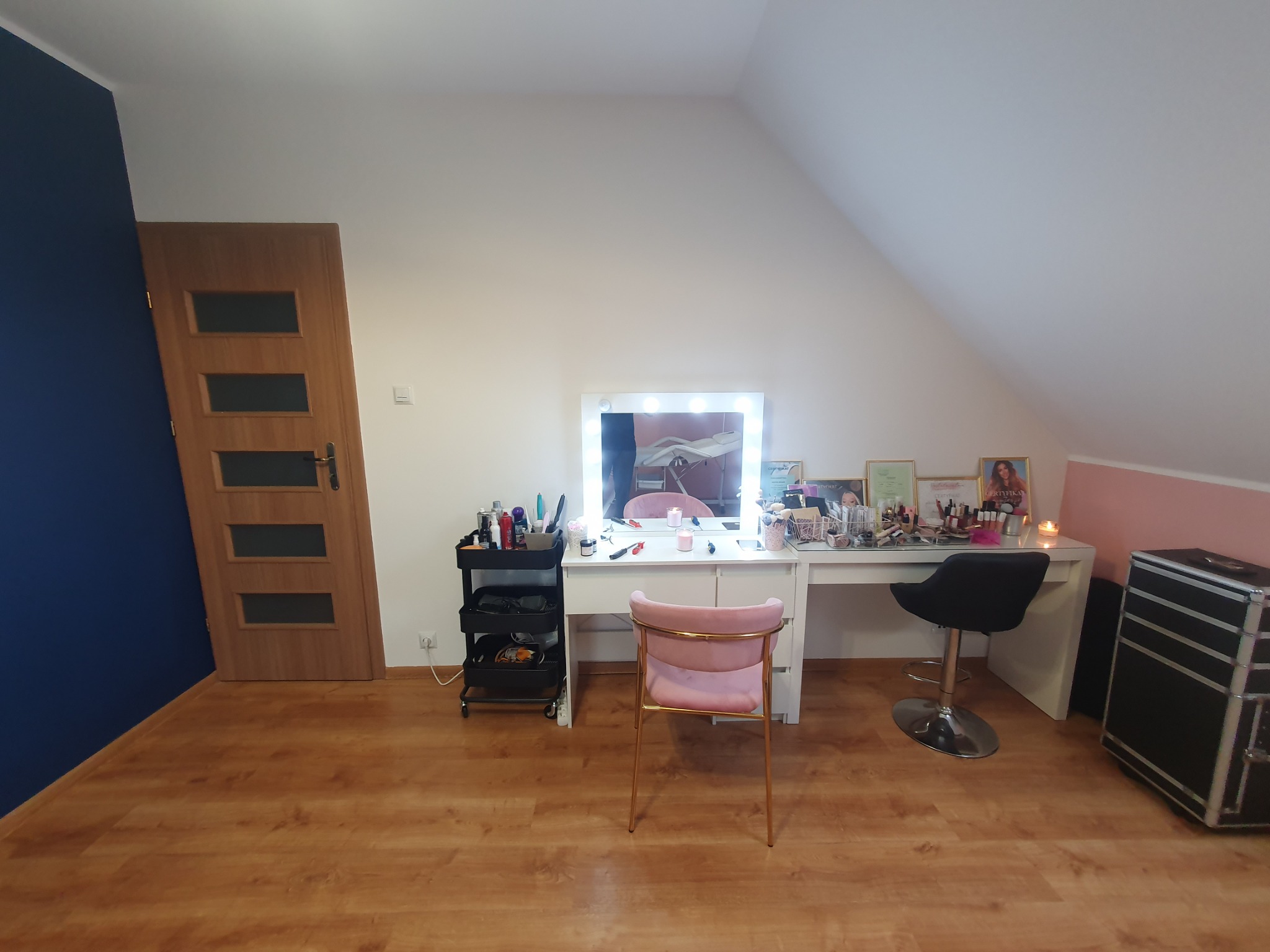 Pokój na poddaszu ze stanowiskiem do makijażu: biała toaletka z oświetleniem LED, różowe krzesło, czarny taboret obrotowy, wózek z kosmetykami, kufer kosmetyczny. Na podłodze panele imitujące...