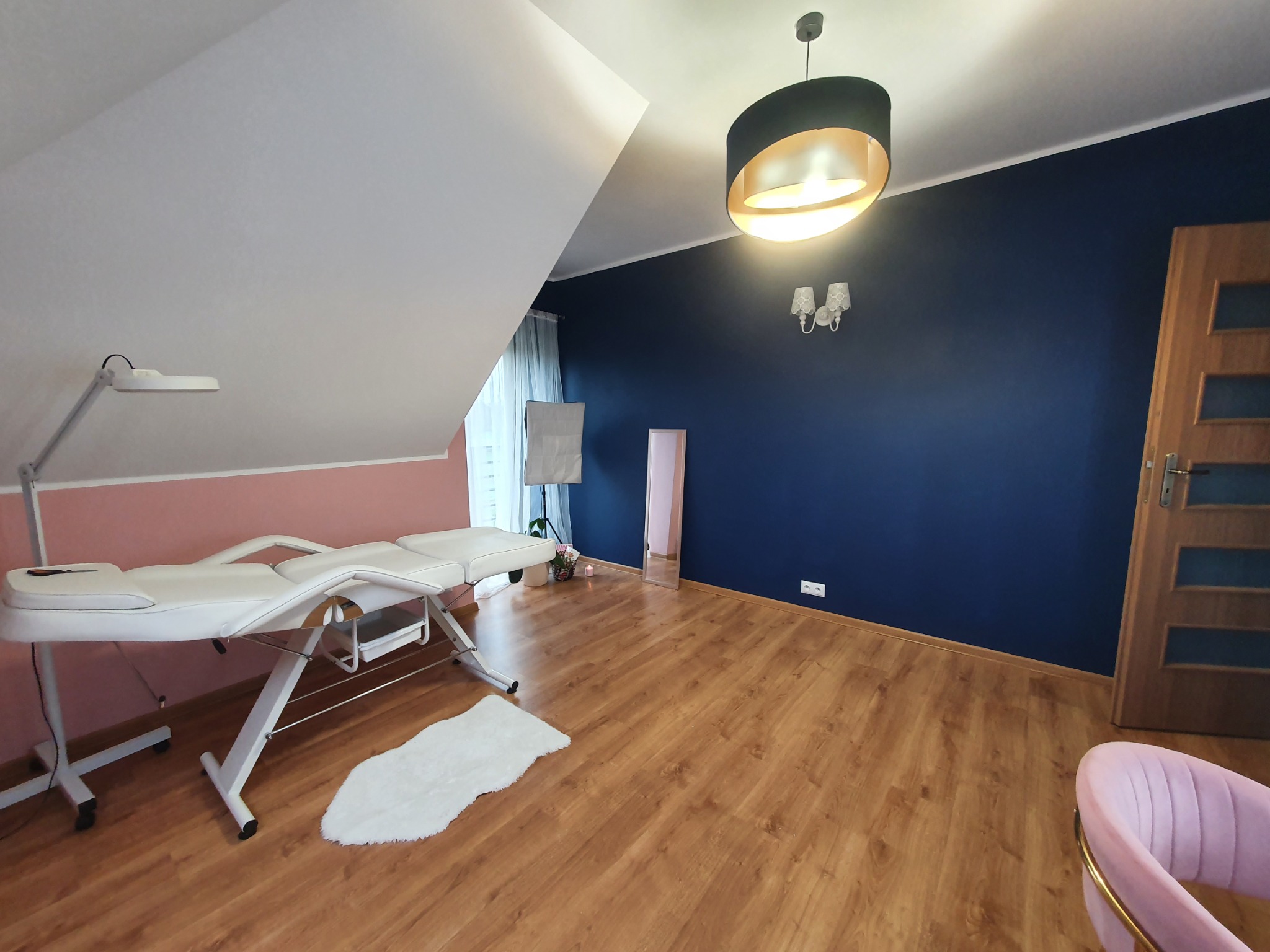 Jasne wnętrze gabinetu kosmetycznego na poddaszu z białym fotelem do zabiegów, drewnianą podłogą i ciemnoniebieską ścianą z ozdobnym oświetleniem.