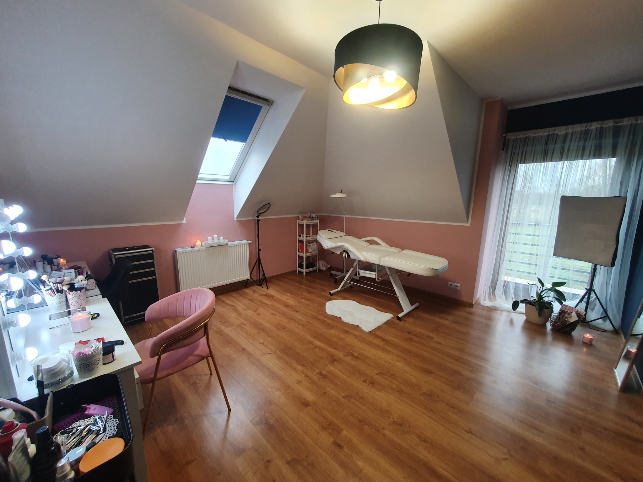 Jasne pomieszczenie na poddaszu zaaranżowane jako gabinet kosmetyczny z drewnianą podłogą, białymi ścianami z różowym pasem, oknem dachowym, lampą wiszącą, łóżkiem kosmetycznym, toaletką...