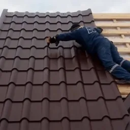 Pracownik w kombinezonie montuje brązowe blachodachówki na drewnianej konstrukcji dachu, widoczna wieża telekomunikacyjna w tle.