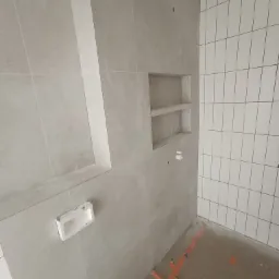 Wnęka z półkami w szarej płytce oraz fragment ściany z białych płytek ułożonych pionowo podczas remontu łazienki.