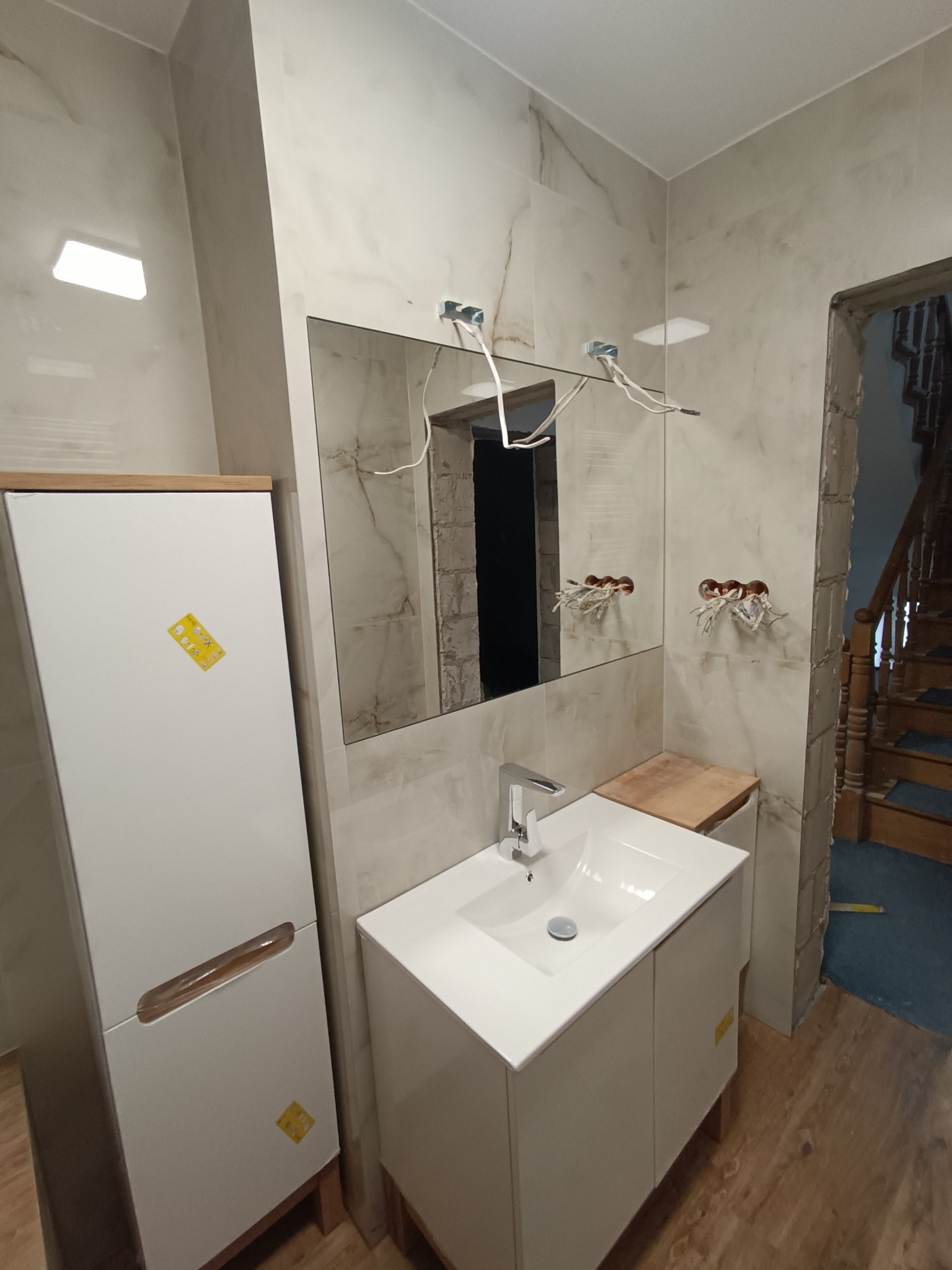 Biała szafka łazienkowa z umywalką i lustrem w trakcie montażu, widoczne wystające kable elektryczne i surowe ściany, drewniane schody w tle.