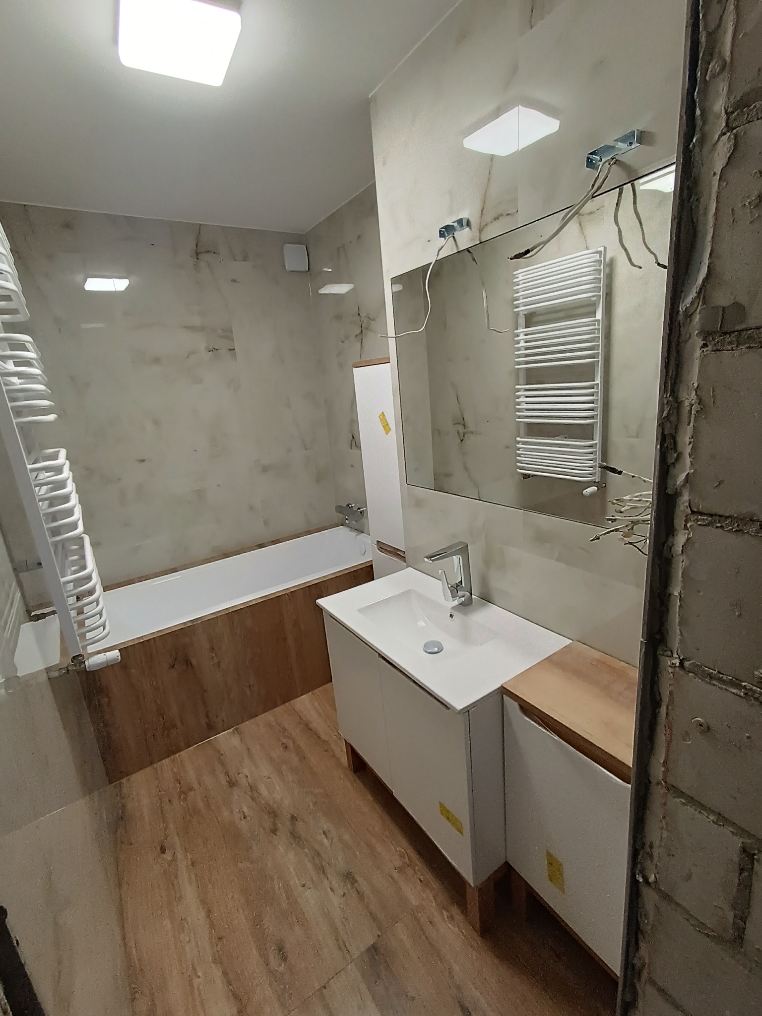 Wnętrze łazienki w trakcie prac wykończeniowych, widoczne płytki imitujące marmur, biała umywalka z szafką, wanna obudowana drewnopodobną płytką oraz fragment surowej ściany z cegły.