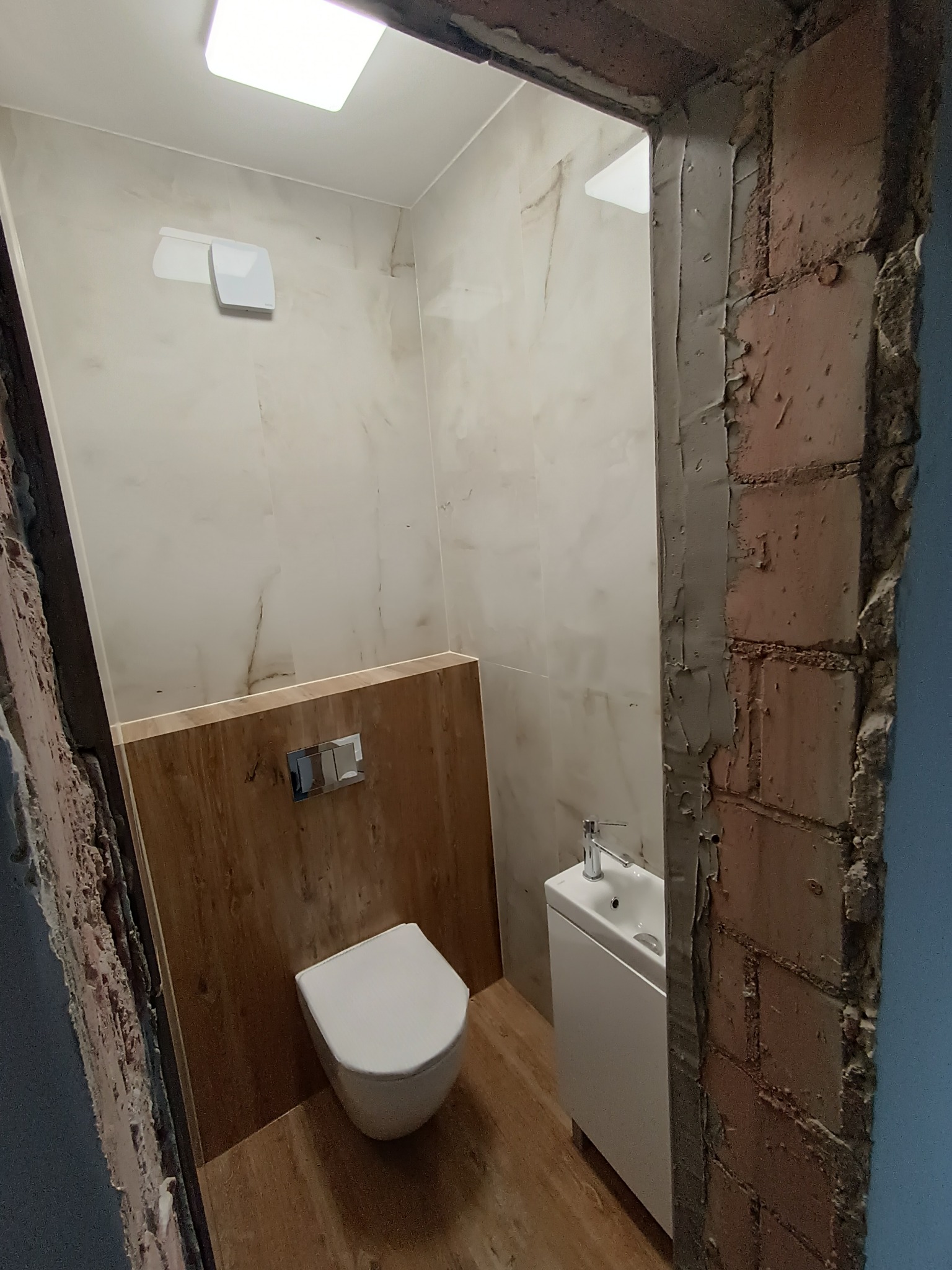 Nowoczesna toaleta w trakcie remontu w Poznaniu, widok zza surowej, otynkowanej framugi drzwi, ukazujący białą ceramikę i panele imitujące drewno.