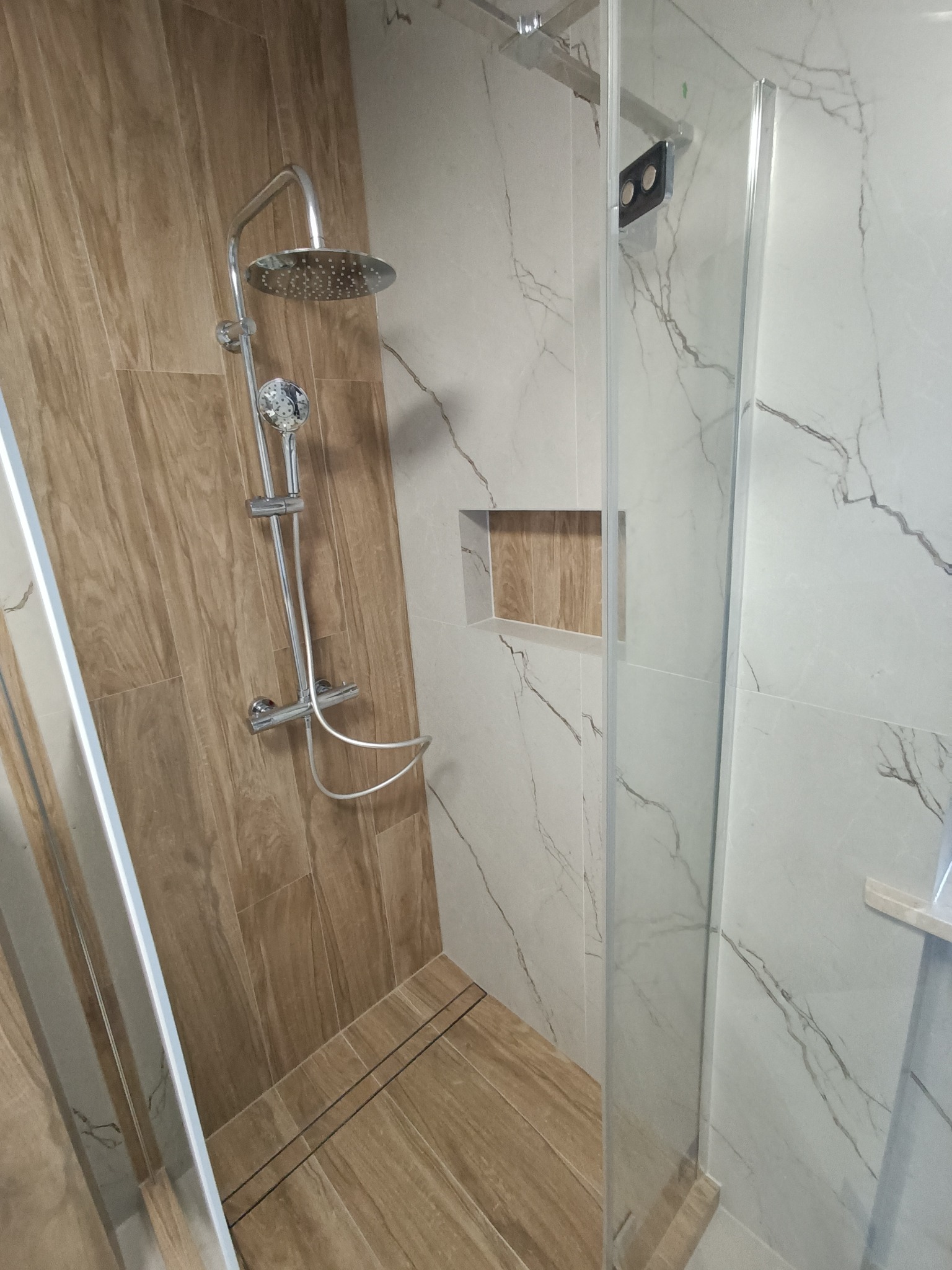 Nowoczesna kabina prysznicowa z deszczownicą, wykończona płytkami imitującymi drewno i marmur z szarą żyłą, z wnęką w ścianie wyłożoną drewnem.