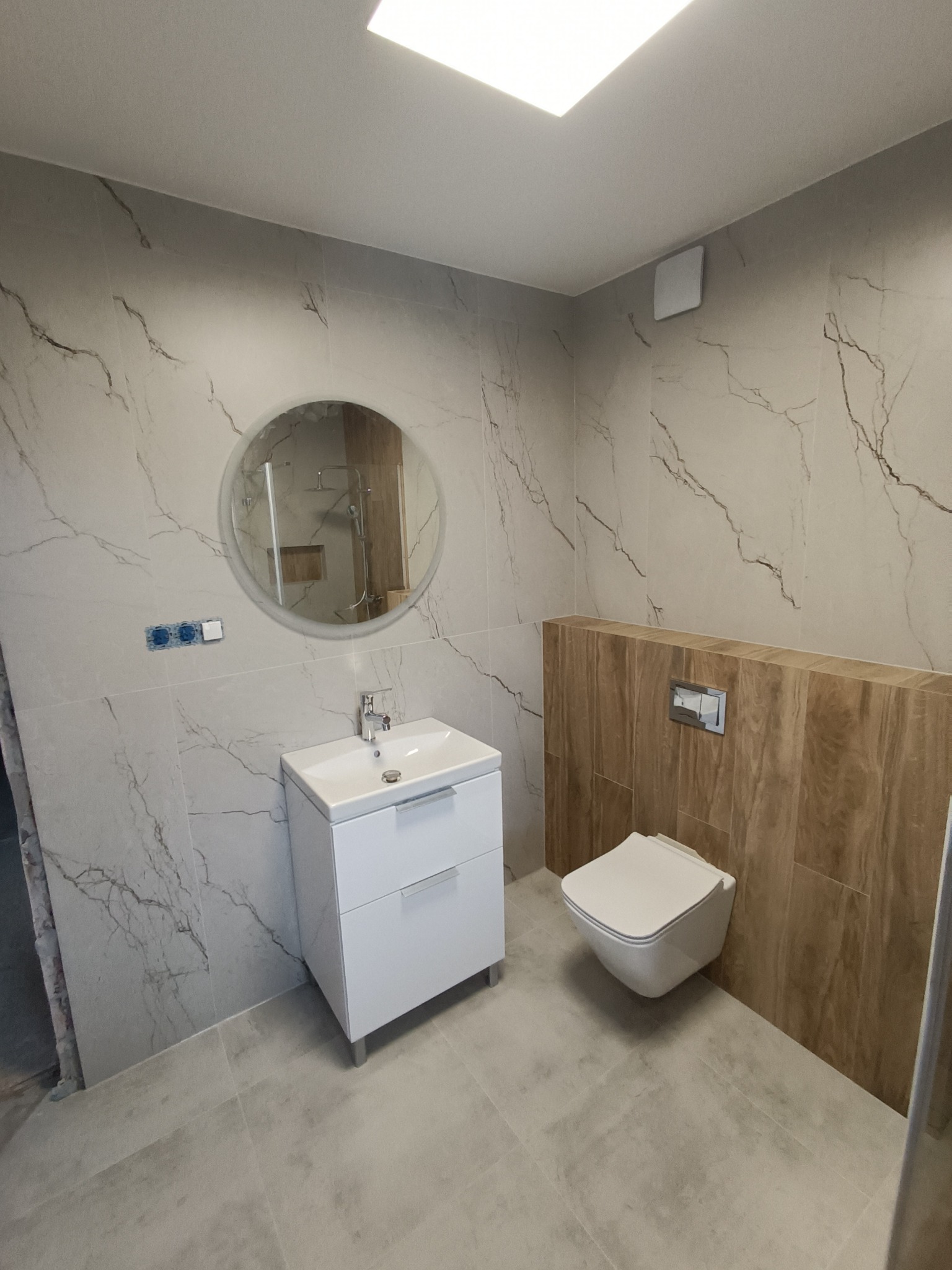 Nowoczesna łazienka z umywalką na szafce, okrągłym lustrem i podwieszaną toaletą na tle marmurowych i drewnopodobnych płytek.