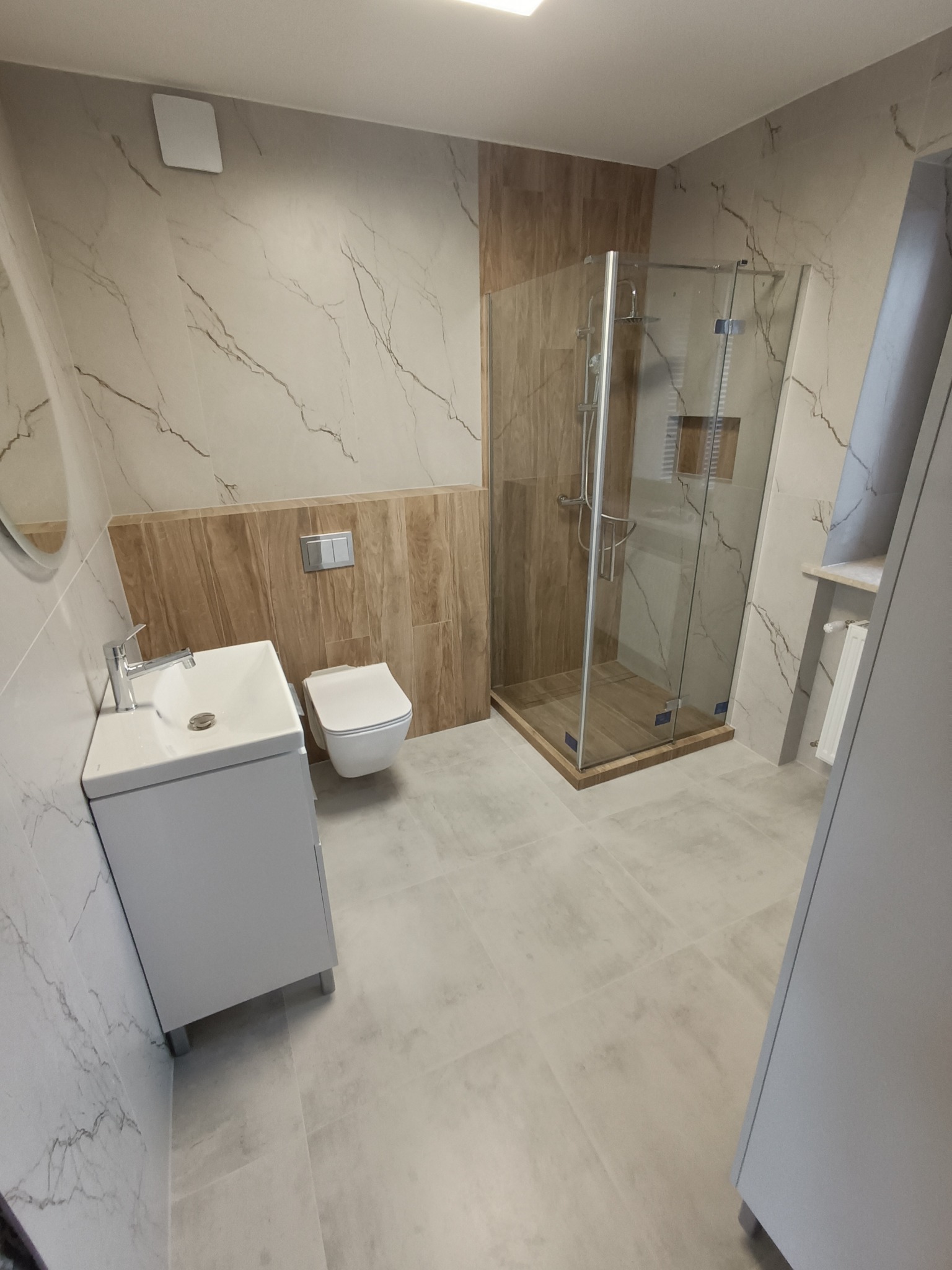 Nowoczesna łazienka z kabiną prysznicową typu walk-in, umywalką nablatową na szafce, wiszącą toaletą i płytkami imitującymi marmur na ścianach.