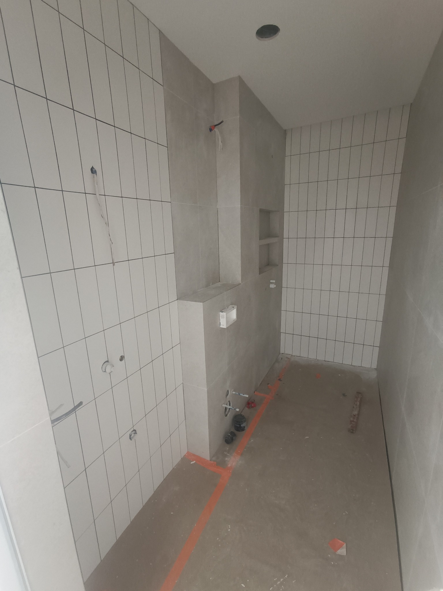 Wnętrze łazienki w trakcie układania płytek ściennych w odcieniach szarości, widoczne przygotowane instalacje, wyznaczone linie na podłodze i wnęka w ścianie.