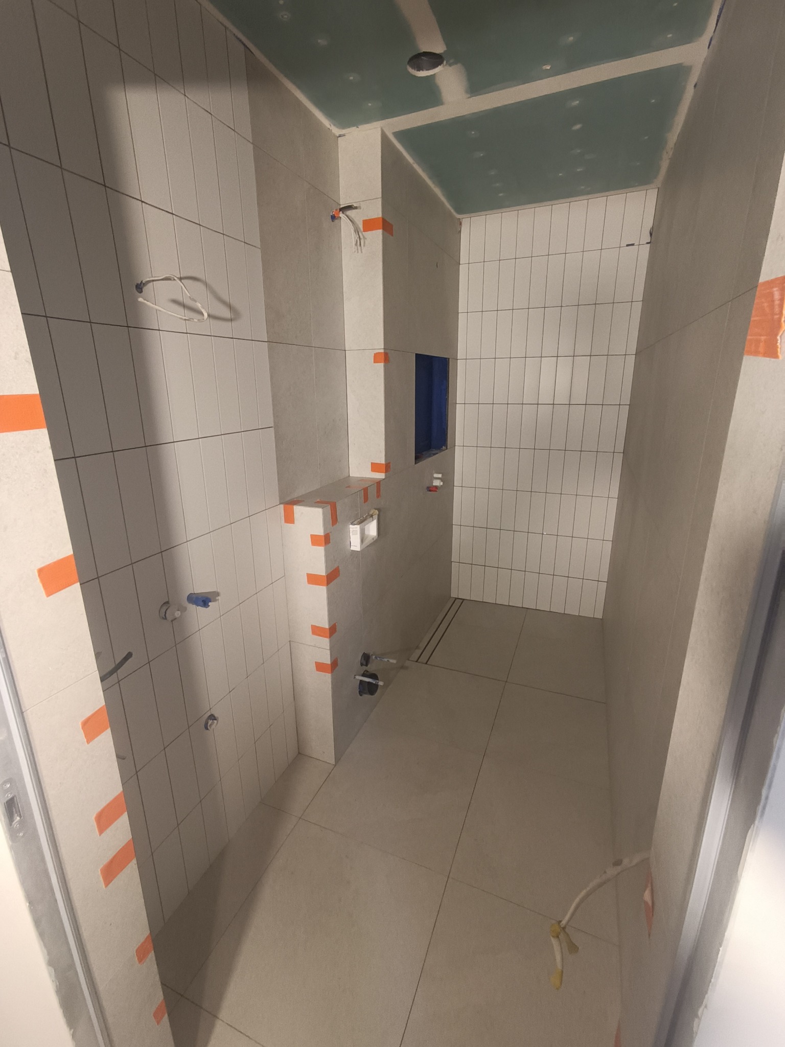 Wnętrze łazienki w trakcie prac wykończeniowych, widoczne płytki ceramiczne na ścianach i podłodze, zabezpieczone taśmą malarską, oraz sufit z płyty kartonowo-gipsowej przed malowaniem. W ścianie...