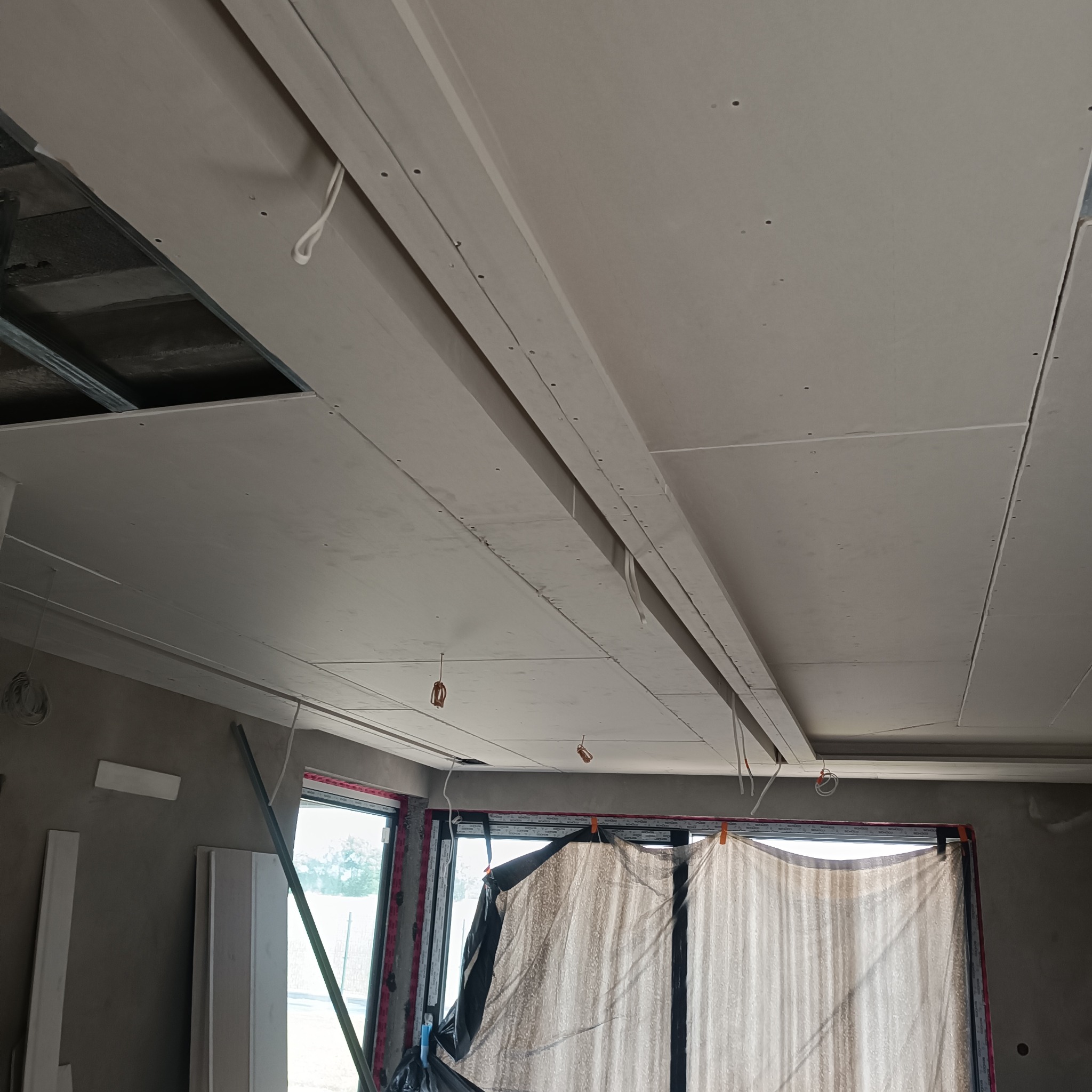 Sufit w trakcie montażu, widoczne płyty gipsowe, metalowa konstrukcja nośna, okno zasłonięte folią ochronną, kable zwisające z sufitu.