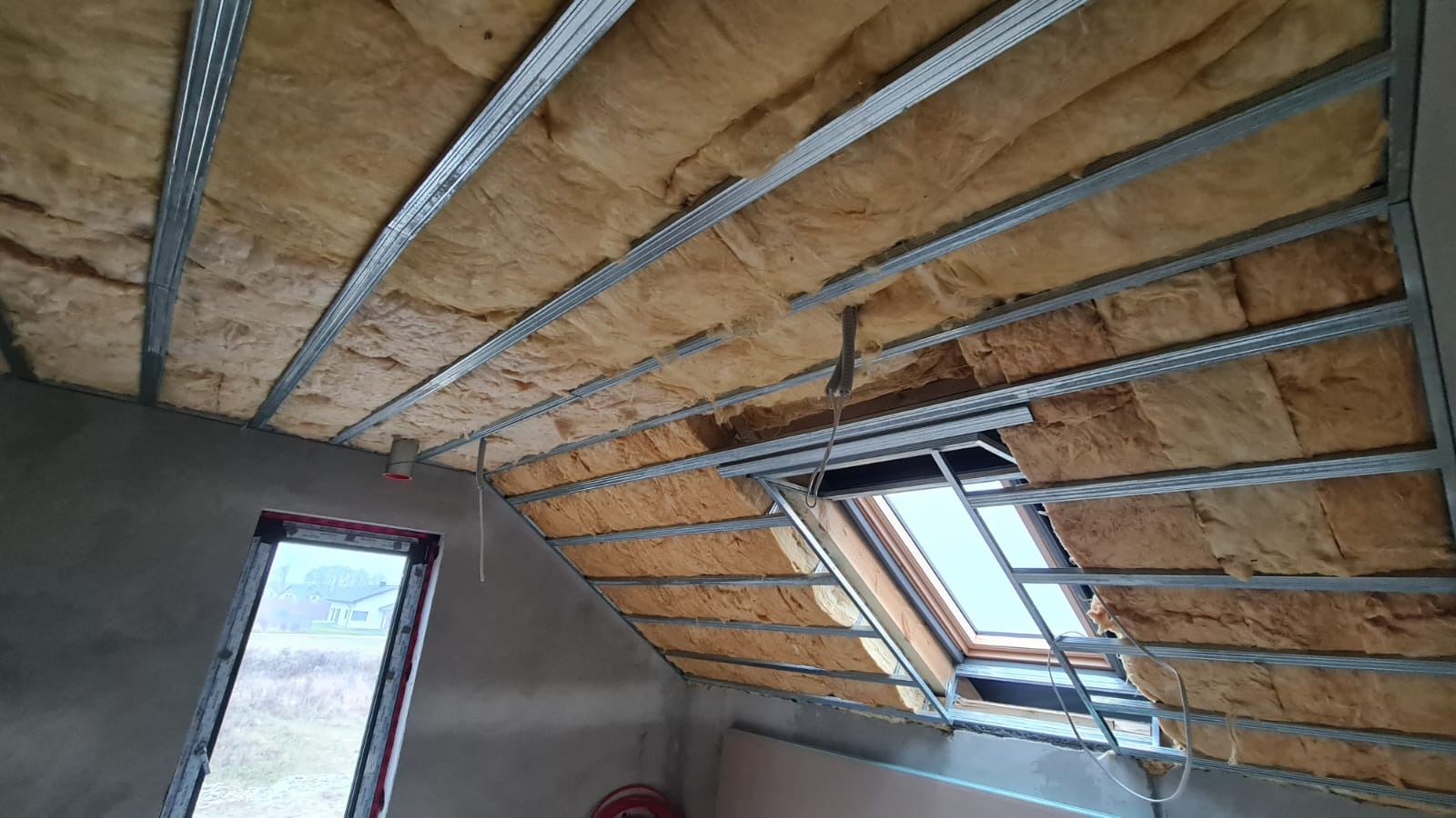 Poddasze z widoczną izolacją termiczną między metalowymi profilami, okno dachowe w trakcie montażu, surowe ściany w tle.