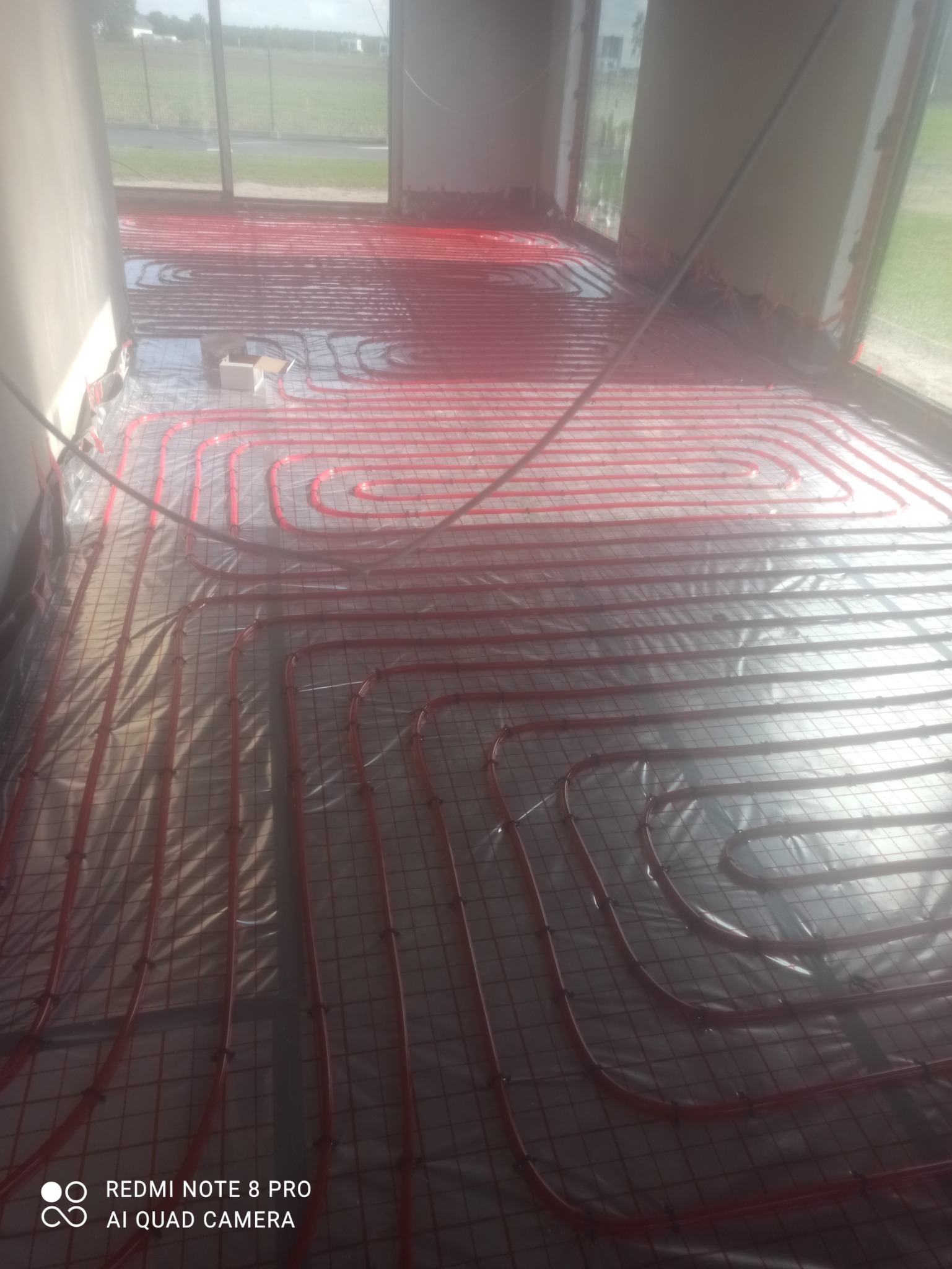 Instalacja ogrzewania podłogowego z czerwonych rurek na folii izolacyjnej, widoczne mocowania i siatka zbrojeniowa, surowy stan wykończenia wnętrza.