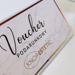Elegancki, marmurkowy voucher prezentowy z czarnym, kaligraficznym napisem 'Voucher' i złotymi elementami, w tym logo 'KNOPESTETIC' i obramowaniem.