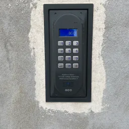 Szary domofon cyfrowy ACO z klawiaturą numeryczną i niebieskim wyświetlaczem, zamontowany na szarej ścianie z widocznymi śladami po niedawnym tynkowaniu.